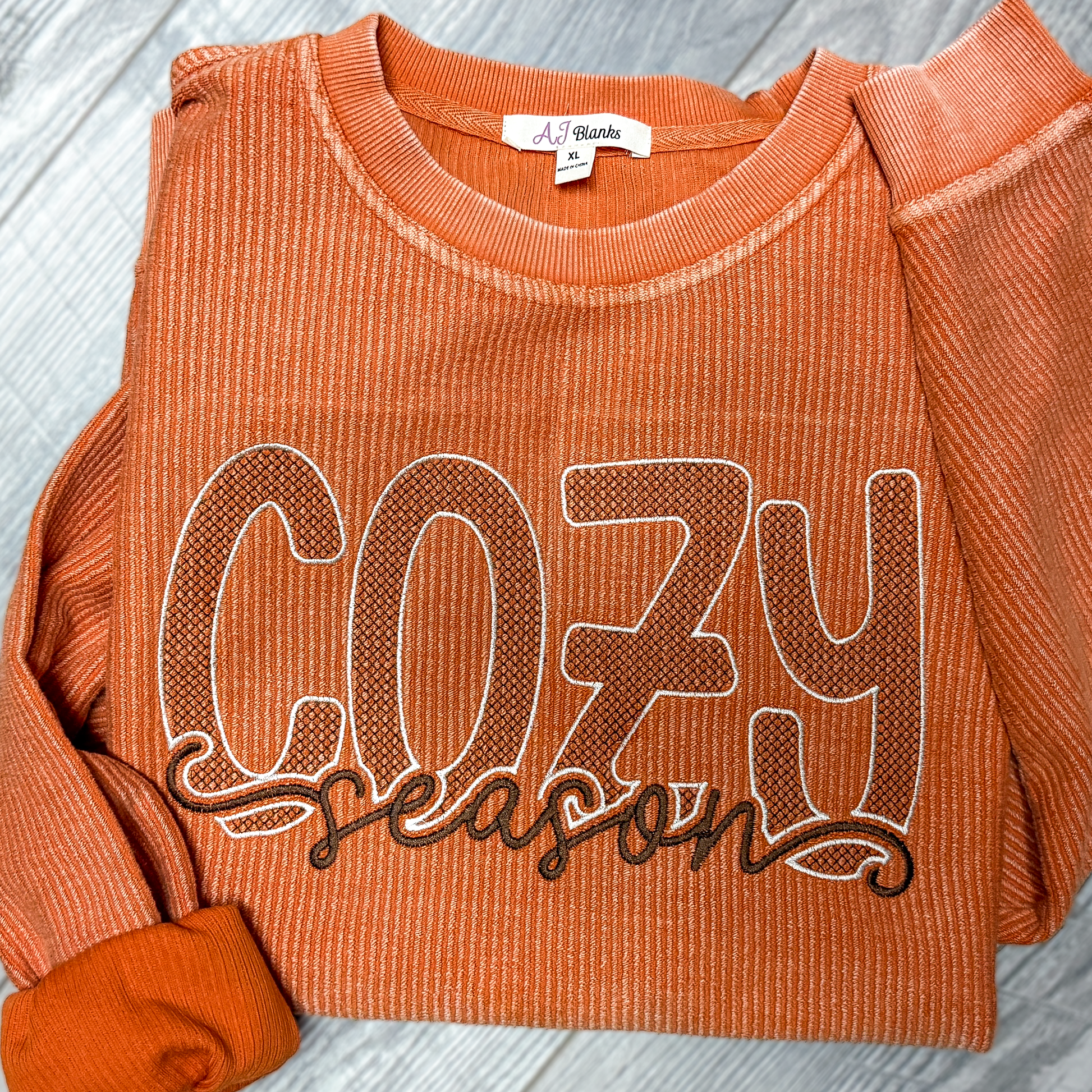 An orange ribbed crewneck sweatshirt with 'COZY season' printed across the front in brown and white, laid flat on a gray wood-textured background.