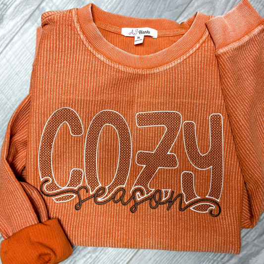 An orange ribbed crewneck sweatshirt with 'COZY season' printed across the front in brown and white, laid flat on a gray wood-textured background.